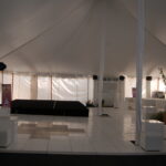 Experience the finest event setting in Atlanta and North Georgia with our spacious tent venue. Located conveniently for residents of Forsyth County, Dawsonville, Dahlonega, and Marietta, our venue features an elegant white canopy and a sophisticated tiled floor. The sides are beautifully furnished with white sofas and tables to provide comfort to your guests. At the front stands a small black indoor stage, accented by decorative plants for added charm. With its softly lit interior, this location is perfect for any rental event you are planning in North Georgia or the surrounding areas such as Atlanta, Forsyth County, Dawsonville, Dahlonega, or Marietta.