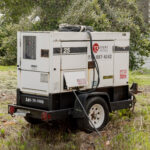 A 20Kw portable white generator with wheels, branded with 25 and T3 Event Rentals, showcases its control panels and cables. Positioned on a grassy area surrounded by trees, it prominently displays a contact number on the side. Perfect for events in Atlanta, this rental from T3 Event Rentals ensures reliable power supply for your special occasion.