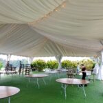 In the scenic locales of Atlanta, North Georgia, Forsyth County, Dawsonville, Dahlonega, and Marietta, a spacious white rental tent liner adorned with string lights provides elegant shelter for a lawn area. The space is beautifully arranged with round and rectangular tables as someone diligently sets up chairs against the lush greenery backdrop.