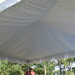 A visitor in a cap enjoys the shade beneath a spacious tent liner ceiling, courtesy of T3 Event Rentals, set up outdoors in Dawsonville. The sunny day highlights the beautiful trees in the background.