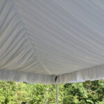 A spacious tent liner canopy featuring elegantly gathered fabric has been set up in a serene outdoor area of Dawsonville. Below the canopy, a bird perches silently as lush green trees rise against the backdrop of a clear sky. All elements, including the tent liner and other event essentials, are available for rent through T3 Event Rentals. Specializing in creating memorable events across Dawsonville and beyond, T3 Event Rentals is your go-to source for quality event rentals.