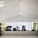 Experience the elegance of this sophisticated rental marquee in North Georgia, perfect for events in Atlanta, Forsyth County, Dawsonville, Dahlonega, and Marietta. Beautifully adorned with chandeliers and white tent liners, it creates a magical atmosphere enhanced by twinkling string lights. The venue includes a fully equipped stage with a drum kit, keyboard, and microphone stands. As sunlight gently filters through the sheer curtains, your events ambiance will be truly unforgettable in these exquisite locations.