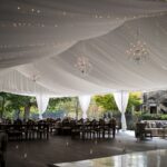 The event space in Atlanta, with its stunning tent liners showcasing a white draped ceiling and elegant chandeliers, is an ideal location for gatherings in North Georgia. In Forsyth County, this venue stands out with neatly arranged rows of tables and chairs, enhanced by soft lighting that creates a cozy feel. Situated conveniently near Dawsonville and Dahlonega, the setting features large windows offering breathtaking views of lush greenery outside. This Marietta-area event venue promises both charm and accessibility for any occasion.