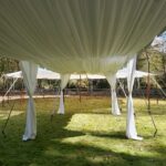 Elegant tent liners gracefully adorn a spacious outdoor rental tent situated on lush green grass, supported by sturdy metal poles. The beautiful backdrop of trees enhances the picturesque setting under the sunny sky. Discover this captivating scene in prime locations such as Atlanta, North Georgia, Forsyth County, Dawsonville, Dahlonega, and Marietta.