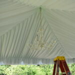 Enhance your event in Dahlonega with a spacious tent featuring elegant white Tent Liners and a vintage crystal chandelier as the centerpiece, available through T3 Event Rentals. The airy design offers open views of lush green trees, perfect for any outdoor celebration. A convenient yellow stepladder is included for setup adjustments. Remember, all items you see are for rent from T3 Event Rentals, your trusted source in the North Georgia area.