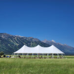 A spacious 20 Rope And Pole Tent from T3 Event Rentals graces an open grassy field in Dahlonega under a clear blue sky, with majestic snow-dappled mountains in the distance. Perfect for events throughout North Georgia.
