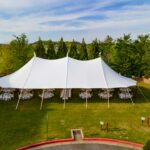 A beautiful Sailcloth Rope and Pole Tent from T3 Event Rentals is perfectly set up on a lush green lawn in Dahlonega, surrounded by towering trees and blue skies. Inside the rental tent, round tables and elegant chairs provide comfortable seating for your event guests. All items—including the tent, tables, and chairs—are available to rent from T3 Event Rentals for any special occasion in Dahlonega.