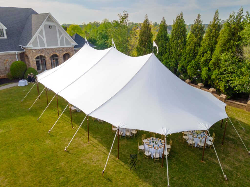 A beautiful sailcloth rope and pole tent from T3 Event Rentals is installed on a lush lawn outside a brick house in Forsyth County. Underneath the elegant rental tent, round tables and chairs are set up for your special event, perfect for gatherings surrounded by tall trees and natural greenery. All tents, tables, and chairs you see here are available for rent exclusively through T3 Event Rentals—serving Forsyth County with top-quality party rentals to make your outdoor event unforgettable.
