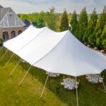A beautiful sailcloth rope and pole tent from T3 Event Rentals is installed on a lush lawn outside a brick house in Forsyth County. Underneath the elegant rental tent, round tables and chairs are set up for your special event, perfect for gatherings surrounded by tall trees and natural greenery. All tents, tables, and chairs you see here are available for rent exclusively through T3 Event Rentals—serving Forsyth County with top-quality party rentals to make your outdoor event unforgettable.