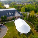A beautiful Sailcloth Rope and Pole Tent from T3 Event Rentals is set up on a lush lawn near a stone building, with green trees, a small parking lot, and a scenic lake in the background—perfect for outdoor events in Forsyth County. All tents, tables, and event rentals are provided by T3 Event Rentals for your next Forsyth County celebration.