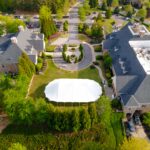 Aerial view of a large Sailcloth Rope and Pole Tent rental set up by T3 Event Rentals on a green lawn between two stone buildings in Dahlonega, surrounded by landscaped paths, mature trees, parked cars, and guests gathering in the courtyard. Perfect for events in the Dahlonega area—tent and event rentals provided by T3 Event Rentals.