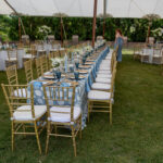 A long table with gold chairs, elegant blue and white patterned tablecloths, vibrant floral centerpieces, and glassware is arranged outdoors beneath a Sailcloth Rope and Pole Tent in Forsyth County. All items are high-quality event rentals provided by T3 Event Rentals. A guest in a blue dress stands at the end of the beautifully set table. Elevate your outdoor celebration in Forsyth County with tent and party rentals from T3 Event Rentals.
