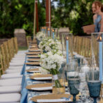 Certainly! Here’s a location-optimized rewrite for the Forsyth County area, based on your requirements: In Forsyth County, an elegant outdoor table set from T3 Event Rentals showcases gold chairs, blue candles and napkins, white floral centerpieces, and assorted glassware beneath stunning Sailcloth Rope and Pole Tents. Two people are visible in the background. All table settings, décor pieces, tents, and seating are rentals provided by T3 Event Rentals for special occasions in Forsyth County.