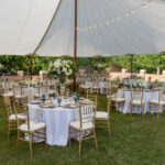 Experience a stunning outdoor event in Forsyth County under a beautiful Sailcloth Rope and Pole Tent from T3 Event Rentals. This setup features round tables dressed in white linens, gold chairs, elegant place settings, floral centerpieces, and twinkling string lights overhead. All rental items are provided by T3 Event Rentals to create an unforgettable atmosphere on grassy grounds with trees in the background. Perfect for weddings or special occasions in Forsyth County!.