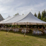 A beautiful sailcloth rope and pole tent with a stunning peaked roof is installed on grass in the heart of Dahlonega, perfect for your next outdoor event. Beneath the tent, T3 Event Rentals provides elegant round tables and chairs surrounded by charming string lights. The scene is set near a building with scenic trees and clear skies in the background. All items are available to rent through T3 Event Rentals—your trusted rental company serving Dahlonega and the surrounding region.