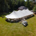 A stunning Sailcloth Rope and Pole Tent from T3 Event Rentals is beautifully set up on a lush grassy lawn in Dawsonville, surrounded by mature trees. Underneath the tent, our rental tables and chairs create an inviting space for your next event. A smoker or grill—available as a rental—adds ambiance with gentle smoke wafting near the tent entrance. Trust T3 Event Rentals for all your Dawsonville party and event rental needs!.