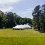 A Sailcloth Rope and Pole Tent from T3 Event Rentals is beautifully set up on a grassy field among tall trees in Dawsonville, with a few guests and rental vehicles nearby under sunny skies. For premier tent rentals and event setups in Dawsonville, trust T3 Event Rentals.