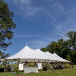 A beautiful Sailcloth Rope and Pole Tent from T3 Event Rentals is set up outdoors on lush green grass under a clear blue sky in the heart of Dawsonville. Surrounded by towering trees, this elegant tent features rental tables and stylish decorations both inside and around the space—perfect for outdoor events in Dawsonville. All items pictured are available for rent through T3 Event Rentals, your trusted source for event rentals near Dawsonville.