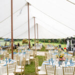 A Sailcloth Rope and Pole Tent by T3 Event Rentals is beautifully set up on a grassy lawn in Dahlonega, featuring round tables draped in white linens, elegant gold chairs, vibrant floral centerpieces, and striking blue glassware. All items are rentals provided by T3 Event Rentals—your premier source for event rentals in Dahlonega.