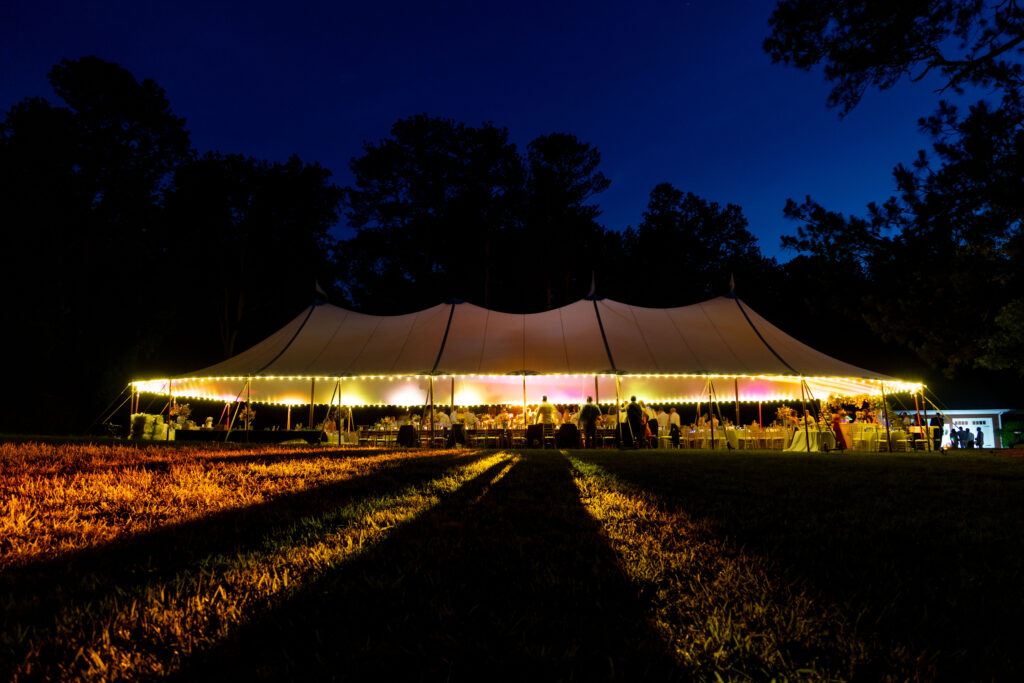 A 44x108' white sailcloth rope and pole tent from T3 Event Rentals glows with string lights at night, casting beautiful shadows on the grass in a wooded area of Forsyth County beneath a deep blue sky. All tents and event items pictured are available to rent from T3 Event Rentals for your next unforgettable Forsyth County celebration.