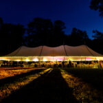 A 44x108' white sailcloth rope and pole tent from T3 Event Rentals glows with string lights at night, casting beautiful shadows on the grass in a wooded area of Forsyth County beneath a deep blue sky. All tents and event items pictured are available to rent from T3 Event Rentals for your next unforgettable Forsyth County celebration.