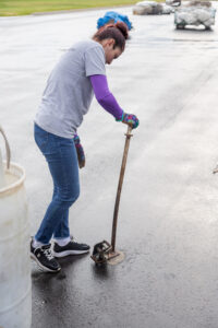 A dedicated individual in a gray shirt and jeans expertly wields a long metal tool on a paved surface, their colorful gloves vividly contrasting against the scene. With their hair neatly tied up, they remain focused amidst the wet pavement thats scattered with wrapped items and a large white container. This is likely part of T3 Event Rentals preparation work for events in Atlanta. All items seen are rentals provided by T3 Event Rentals, specializing in crafting memorable events throughout the region.