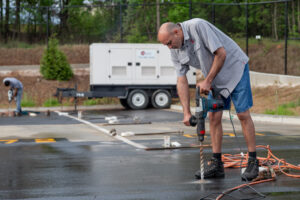 In the heart of Forsyth County, a skilled man in a gray shirt and denim shorts deftly uses a power drill on an outdoor concrete surface, likely setting up for an event with T3 Event Rentals. In the background, another team member diligently works near a trailer and lush trees, emphasizing the spacious ambiance of a parking lot or flat paved area. T3 Event Rentals is your go-to source for all your rental needs in this vibrant region.