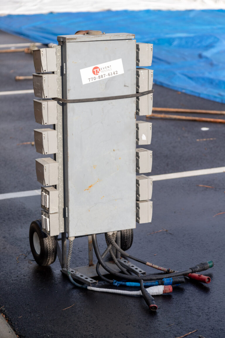 A mobile electrical power distribution box on wheels is stationed on the wet pavement, primed for event rentals in Dawsonville. This unit, available through T3 Event Rentals, boasts multiple outlets along its sides and several cables at the bottom. The front displays a contact label for easy communication. In the background, a blue tarp adds to the scenes setting appeal.