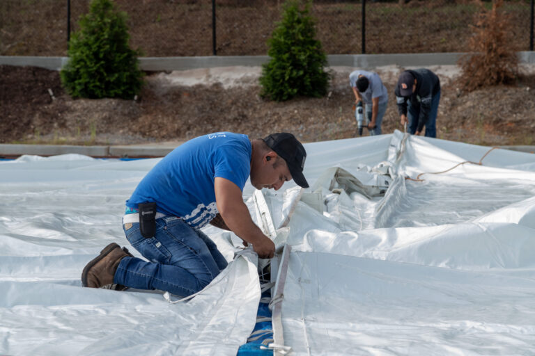 In North Georgia, a man wearing a blue shirt and black cap kneels to secure a large white tarp, commonly used in event setups, on the ground. In the background, two others from T3 Event Rentals assist him amidst greenery and dirt.