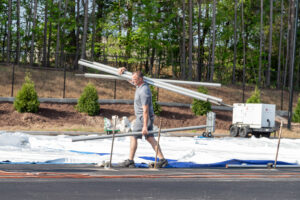 In the city of Atlanta, a man in a gray t-shirt is seen carrying several long metal pipes across a paved outdoor area. He seems to be prepping for an event with T3 Event Rentals. The background features white structures and a portable generator, with trees and shrubbery lining the perimeter.