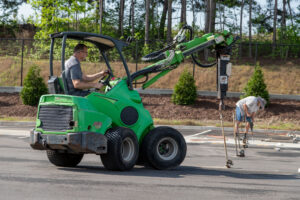 In a bustling parking lot in Dahlonega, a man expertly maneuvers a green mini-loader with a drilling attachment, setting the stage for event rentals. Nearby, someone is diligently working with tools by the fence and trees, preparing the surroundings for an upcoming event. T3 Event Rentals orchestrates these efforts to ensure everything runs smoothly in Dahlonega.