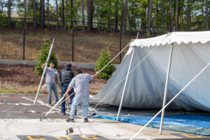 Three men from the T3 Event Rentals team are setting up a large white tent outdoors in Atlanta. They are holding metal poles as part of the tents structure. The scene includes a backdrop of fences and trees, with tarps and parking space markings on the ground.