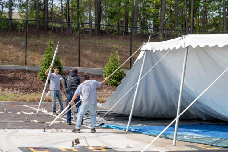Three men from the T3 Event Rentals team are setting up a large white tent outdoors in Atlanta. They are holding metal poles as part of the tents structure. The scene includes a backdrop of fences and trees, with tarps and parking space markings on the ground.
