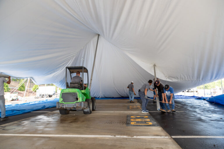 T3 Event Rentals expertly sets up a spacious white tent in a Dawsonville parking lot, with a green utility vehicle conveniently parked inside. A dedicated team of workers from T3 Event Rentals adjusts the tent, which spans across parking spaces uniquely marked with yellow numbers. If youre planning an event in Dawsonville and need reliable rental services, T3 Event Rentals offers exceptional solutions for your event needs.
