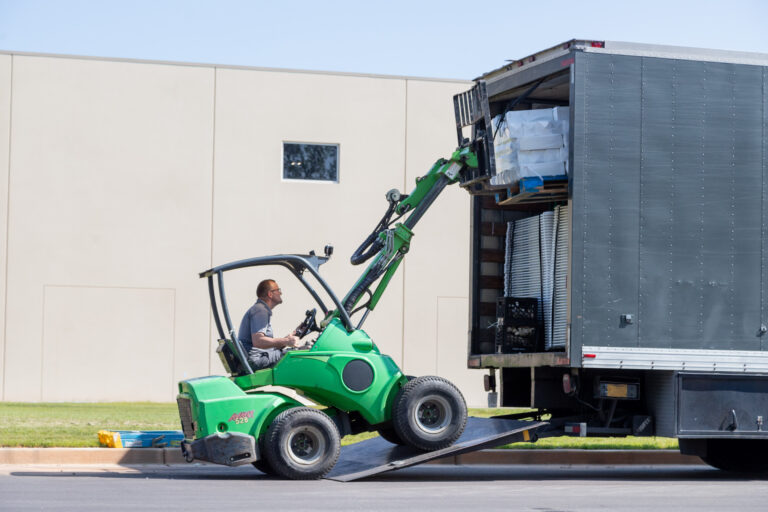 A man operates a green forklift, adeptly lifting rental pallets into the back of a large gray truck parked on a street in Dawsonville. This efficient operation, conducted by T3 Event Rentals, ensures timely delivery for event needs. As the forklift maneuvers up the ramp, the beige building and grass lawn bask under Dawsonvilles clear sky.