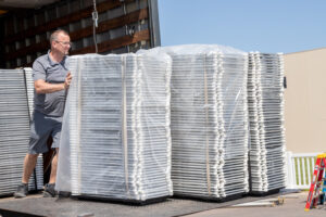 In Forsyth County, a man wearing glasses and a gray shirt is positioned on a truck loading platform next to stacks of large metal frame structures wrapped in plastic, perfect for event rentals. Adding a splash of color to the scene is a red ladder, set against the backdrop of Forsyth Countys clear blue sky. All equipment is available for rent through T3 Event Rentals, your go-to company in Forsyth County for all your event needs.