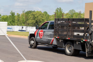 A branded truck from T3 Event Rentals is parked on a paved lot in Forsyth County, featuring a sleek black flatbed with protective side rails. In the background, lush green trees and white portable buildings stand ready to accommodate any event rental needs. Whether youre planning an occasion in Forsyth County or beyond, T3 Event Rentals has you covered.
