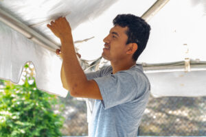 In Forsyth County, a man wearing a gray t-shirt diligently adjusts a white canopy tent, part of the T3 Event Rentals setup. His attention is focused on securing the tent’s edges amidst the clear backdrop of lush greenery and fencing, indicative of a bright sunny day.