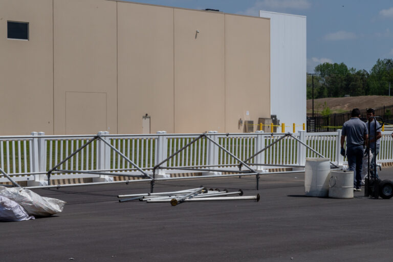 In the heart of Forsyth County, workers are busy preparing a metal structure on an asphalt surface near a beige building accented with white fencing. This setup, potentially for event rentals by T3 Event Rentals, includes an array of white buckets and tools under the area’s famously clear skies. T3 Event Rentals is renowned for transforming spaces across Forsyth County into unforgettable event venues.