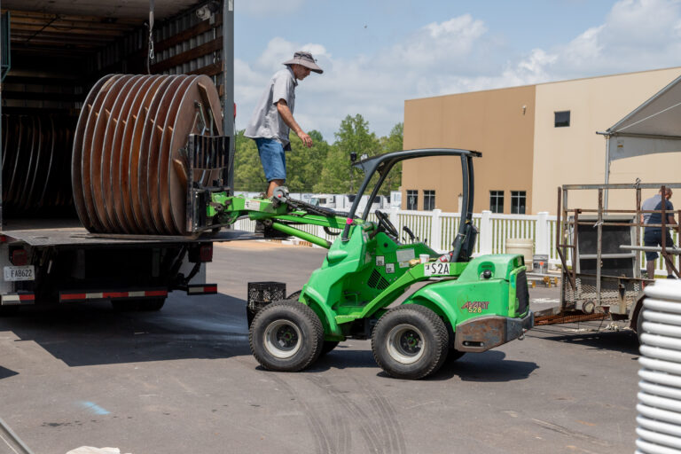 An individual is positioned on a green forklift rented from T3 Event Rentals, adjusting a large coil inside a truck trailer. The scene unfolds outside a beige industrial building in Forsyth County on a sunny day, surrounded by trees and a fence in the background.