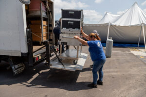 In Atlanta, a person in a blue shirt is seen pushing a cart loaded with T3 Event Rentals equipment onto a trucks lift gate, while another assists from the truck. A large white tent stands in the background, creating an organized setup for an outdoor event on this sunny day. All items showcased by T3 Event Rentals are exclusively for rent.