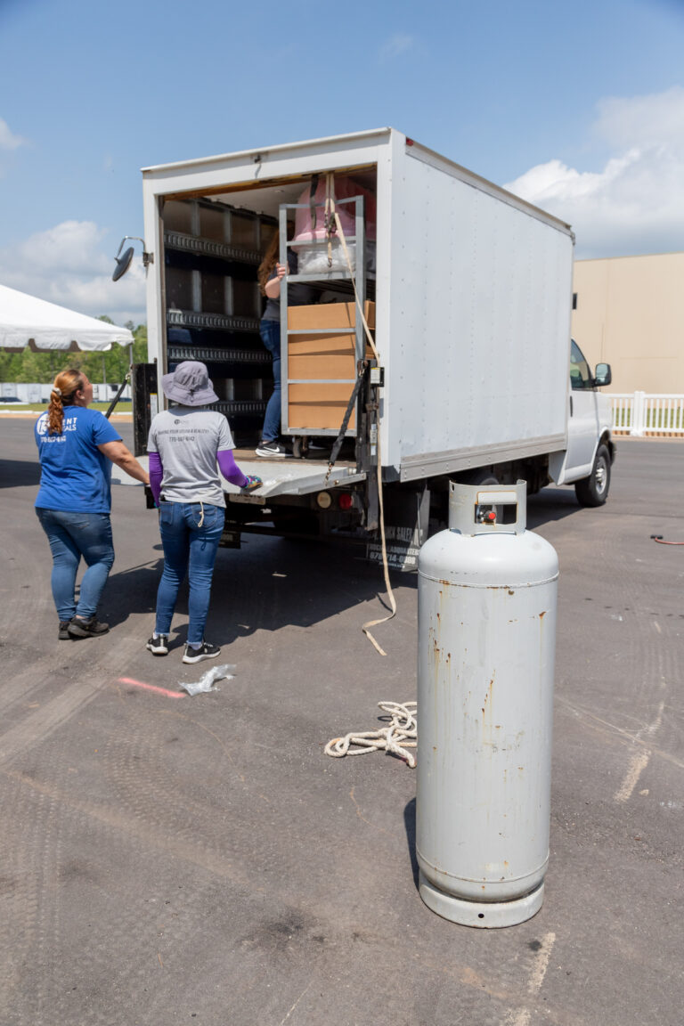 In a Dawsonville parking lot, two individuals are busy loading rental items into the back of a white moving truck from T3 Event Rentals. A large propane tank stands prominently in the foreground as they prepare for another successful event under the clear sky, with just a few clouds scattered above.