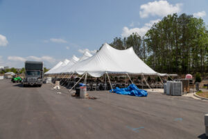 Dahlonega event planners will find the perfect outdoor setup with T3 Event Rentals. Our large white tents, ideal for all your special events, are elegantly arranged in a scenic parking area surrounded by lush trees. Amidst clear skies and a few clouds, our construction materials and equipment—featuring a large truck and signature blue tarp—are meticulously organized to enhance your experience. Trust T3 Event Rentals to transform any location into the event venue of your dreams.