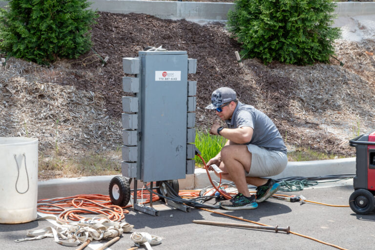 A man crouches on a paved area in Dawsonville beside a T3 Event Rentals setup, working on a large electrical panel with open compartments. Surrounded by orange cables, he wears a cap, sunglasses, blue gloves, and a gray shirt. Trees and mulch provide the backdrop.