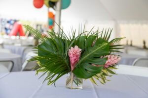 Transform your event in Dawsonville with a breathtaking floral centerpiece from T3 Event Rentals. Featuring vibrant pink flowers and lush tropical leaves, elegantly arranged in a square glass vase, this stunning piece graces any gathering. Set on a white-clothed table under a tent adorned with colorful balloons, our rental displays promise to add charm and elegance. Turn to T3 Event Rentals for all your Dawsonville event needs!.