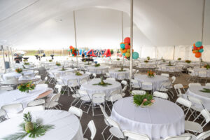Experience a stunning event setup in Dahlonega with T3 Event Rentals. Our expansive tent features rows of white round tables and chairs, each table elegantly dressed with tropical centerpieces. Vibrant balloons create a festive backdrop, perfect for your special occasion. This beautiful arrangement is available through T3 Event Rentals, bringing premier event services to Dahlonega.