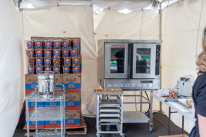 Inside the T3 Event Rentals tent in Forsyth County, a commercial oven quietly hums with trays of delicious food, while boxes filled with bite-sized snacks are neatly stacked on the left. A sturdy metal rack proudly displays a pot and various utensils. To the right, a table overflows with additional items from T3 Event Rentals, and you can spot a person amidst this bustling setup.