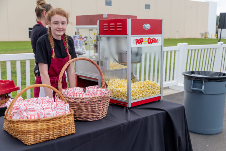 An individual with striking red hair stands beside a T3 Event Rentals popcorn machine and two baskets placed on a table. The baskets are stocked with packets of popcorn. This outdoor scene is set in Dahlonega, featuring a white fence, an expansive building, and a nearby trash can. All items shown are rentals from T3 Event Rentals.