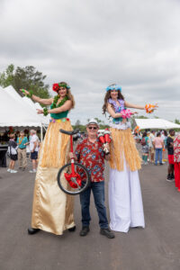 At an outdoor event in Forsyth County, a man in a Hawaiian shirt expertly juggles clubs while balancing on a unicycle, joined by two women on stilts adorned in grass skirts and floral headpieces. This eye-catching display is part of the vibrant entertainment options offered by T3 Event Rentals, perfect for unique occasions. In the background, you can see people milling around and tents adding to the lively atmosphere. Remember T3 Event Rentals for your next unforgettable event experience!.