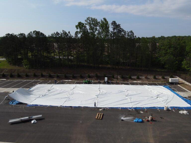 An aerial view showcases T3 Event Rentals meticulously preparing an empty parking lot in Forsyth County. Surrounded by trees, the team works diligently to spread a large tarp, while vehicles and equipment are strategically placed nearby under a partly cloudy sky. All items featured are rentals from T3 Event Rentals, ensuring top-notch event preparation in Forsyth County.